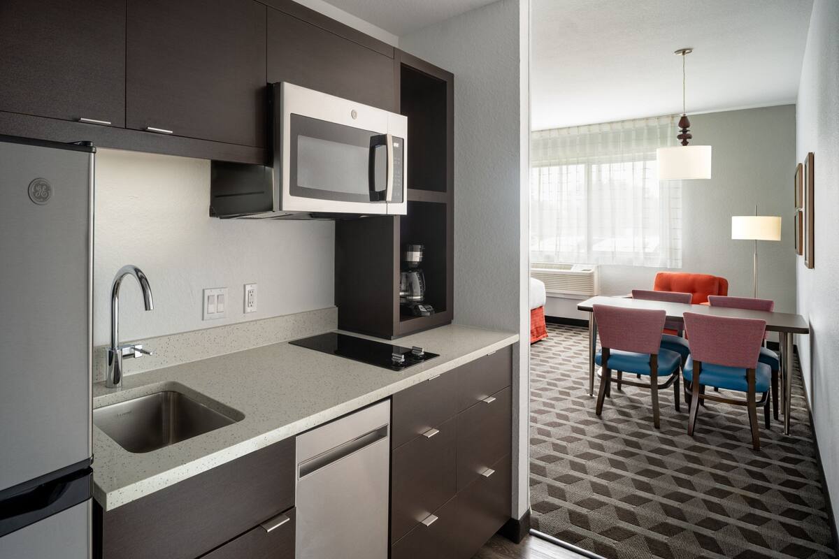 A modern kitchen with a built-in microwave, stainless steel sink, and a coffee machine. There is a dining area with a table and chairs, and a patterned carpet.