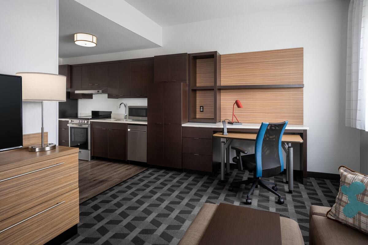 A modern hotel room with a kitchenette, a wooden desk, a chair, a television, and a patterned rug.