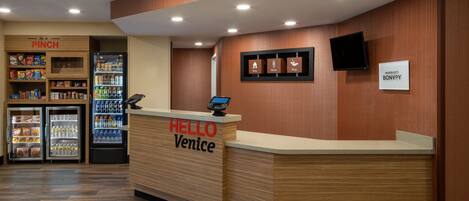 Un espace d’accueil d’hôtel avec un comptoir de réception en bois portant l’inscription « HELLO Venise », un réfrigérateur rempli de boissons et un présentoir « PINCH » proposant des en-cas.