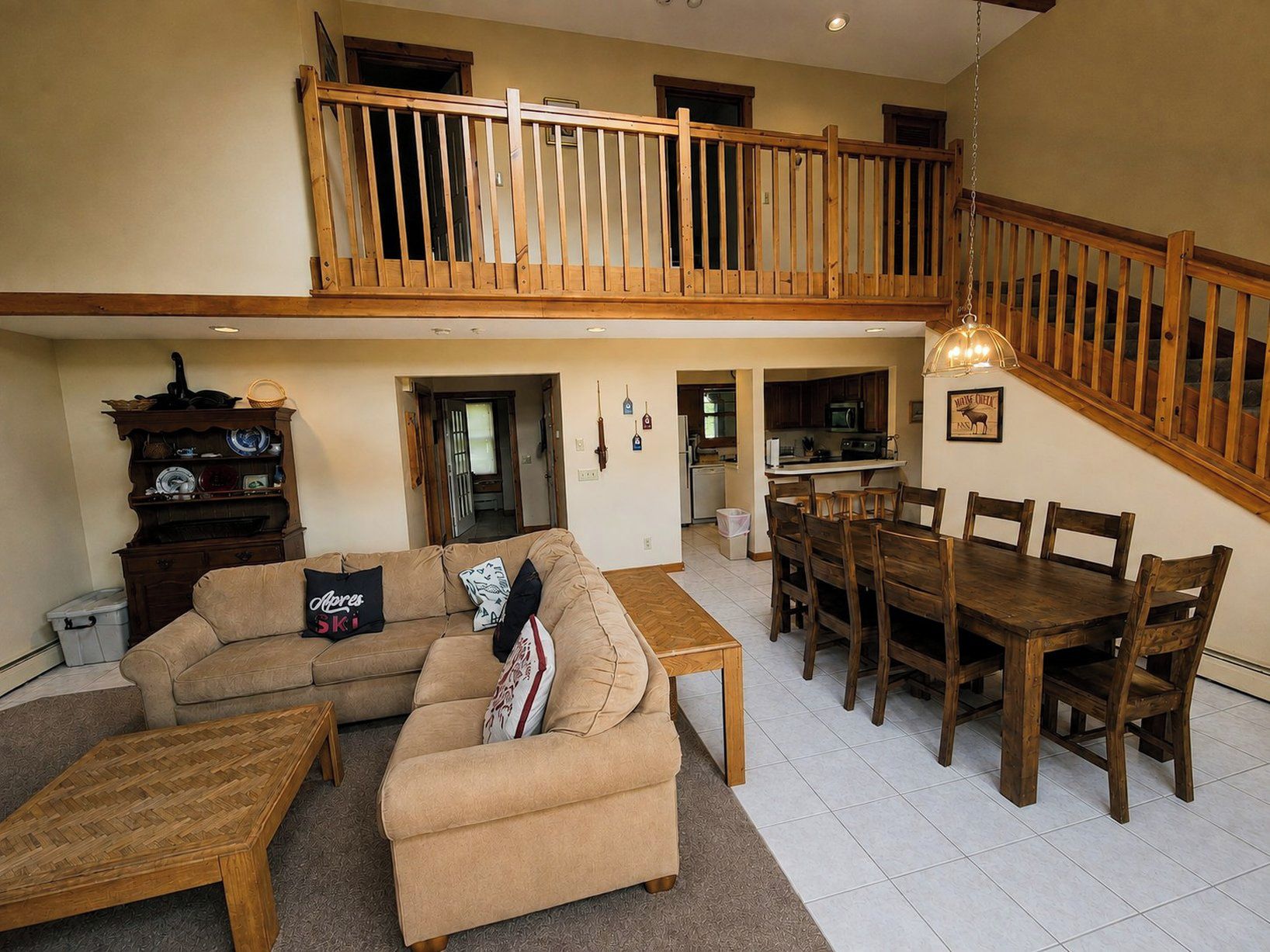 A spacious living area with a high ceiling, a wooden staircase, a dining table with chairs, and a sofa with cushions.