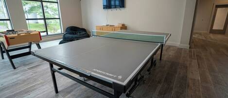 A room with a ping pong table, a foosball table, and a TV mounted on the wall.