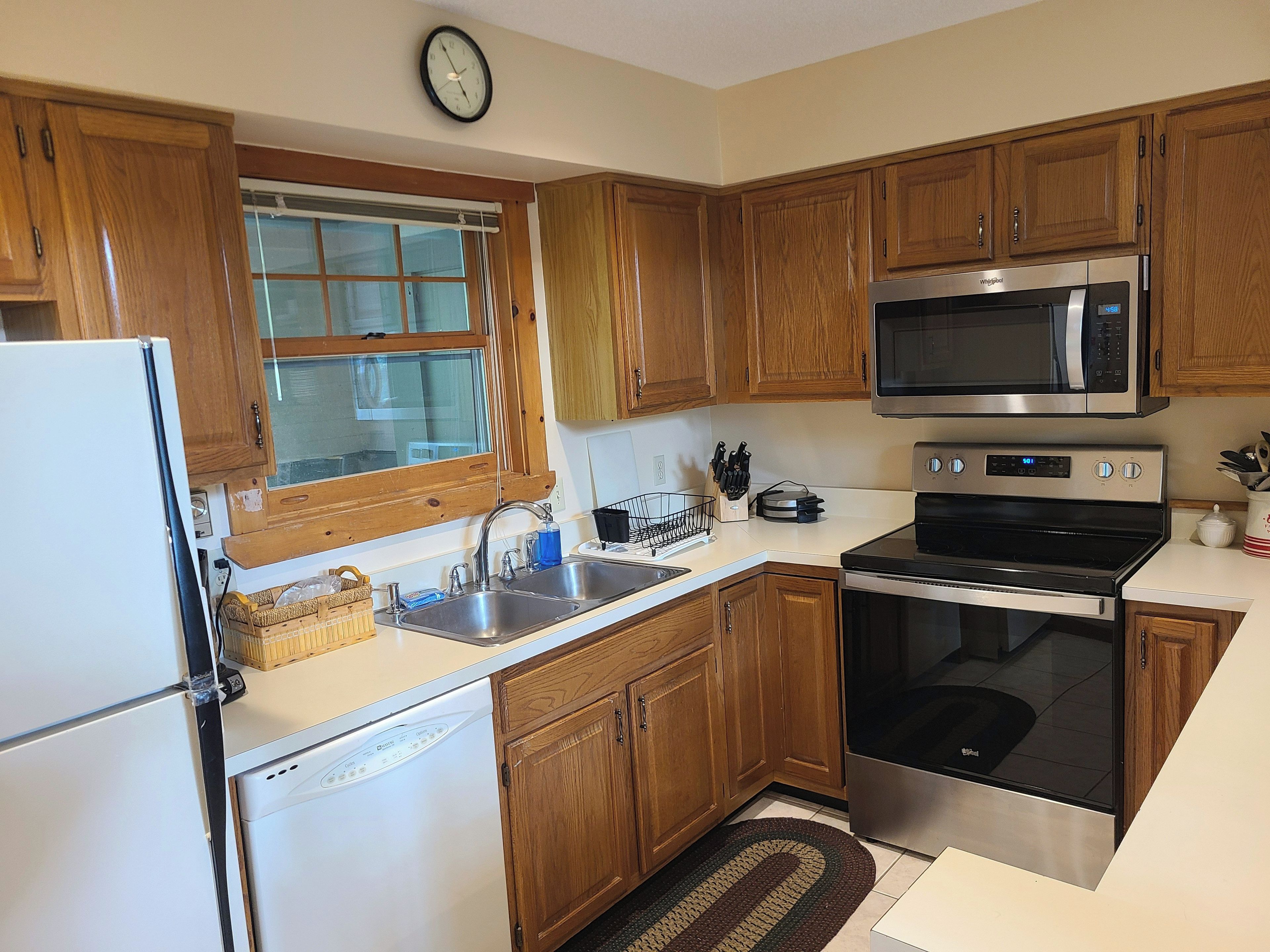 A kitchen with wooden cabinets, a microwave, an oven, a dishwasher, and a window with blinds.
