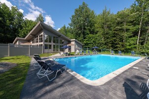 A rectangular swimming pool with a tiled deck, surrounded by lounge chairs and umbrellas, with a building in the background.