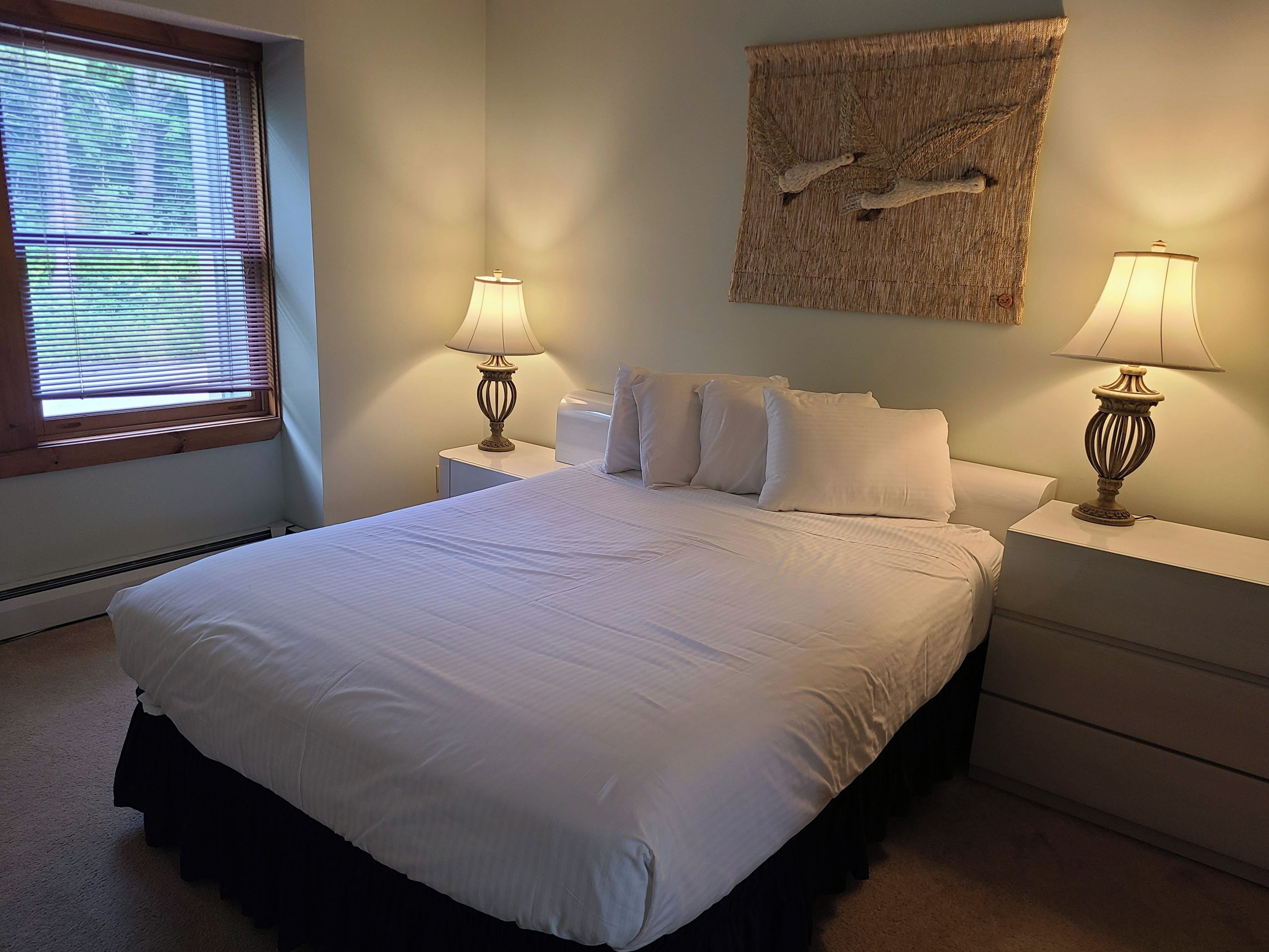 A bedroom with a large bed, two bedside tables with lamps, a window with blinds, and a wall-mounted artwork.