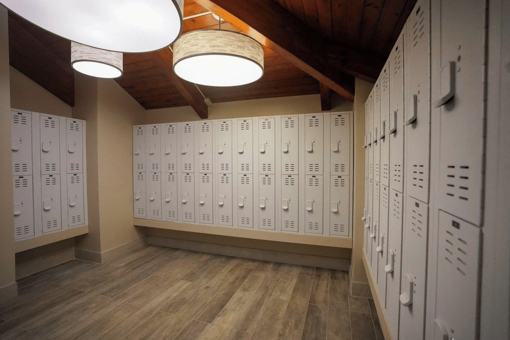 A locker room with multiple white lockers, wooden flooring, and hanging lamps.