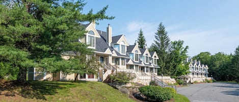 A row of large, multi-story houses with steep roofs and large windows, surrounded by trees and a grassy hill.