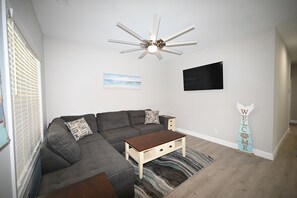 Uma sala de estar moderna com sofá modular, mesa de centro de madeira, ventilador de teto, televisão de tela plana e um letreiro com a palavra 'WELCOME'.