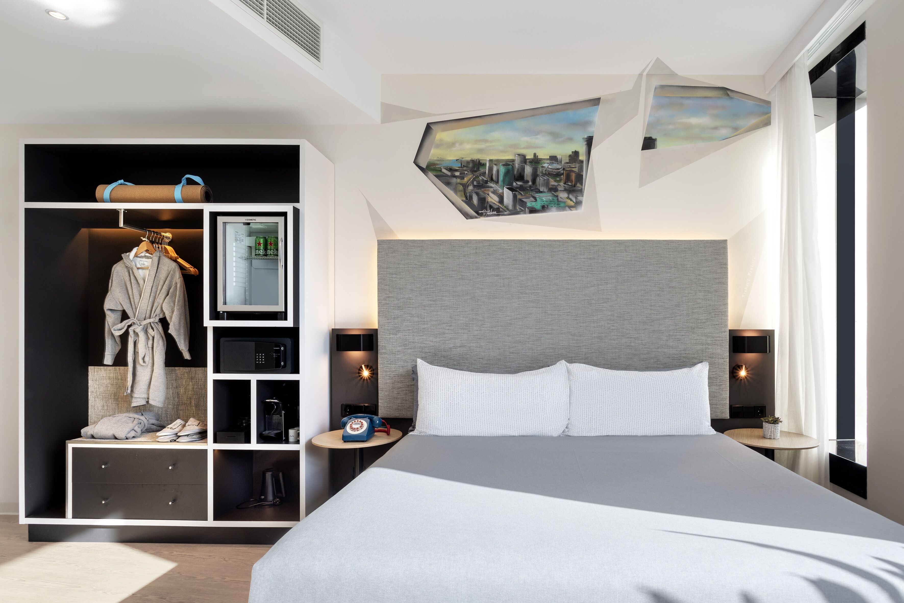 A modern hotel room with a large bed, a built-in wardrobe, a cityscape painting, and a small table with a lamp.