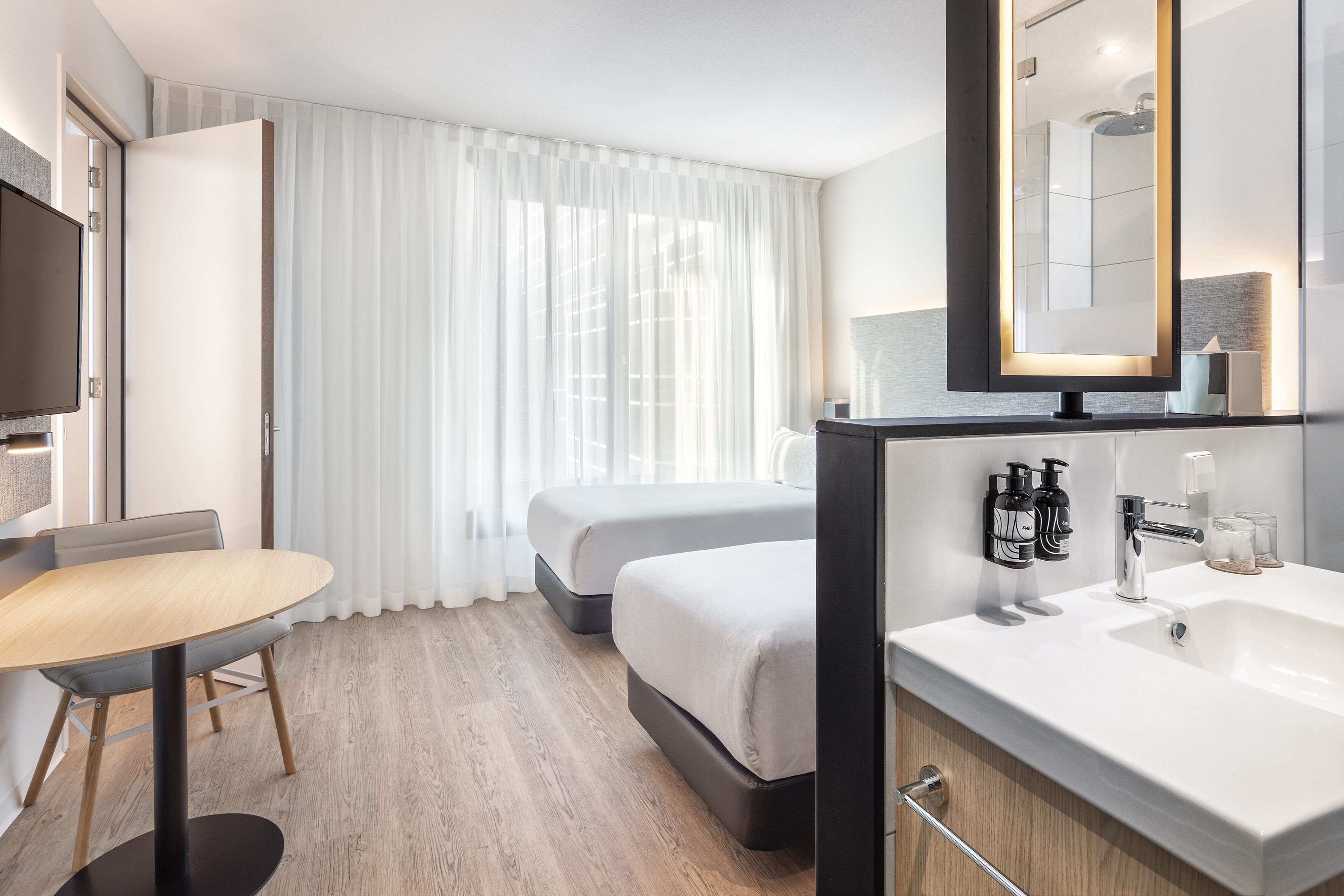 A hotel room with a wooden floor, a bed, a desk with a chair, a TV, a large mirror, and a bathroom with a sink and toiletries.
