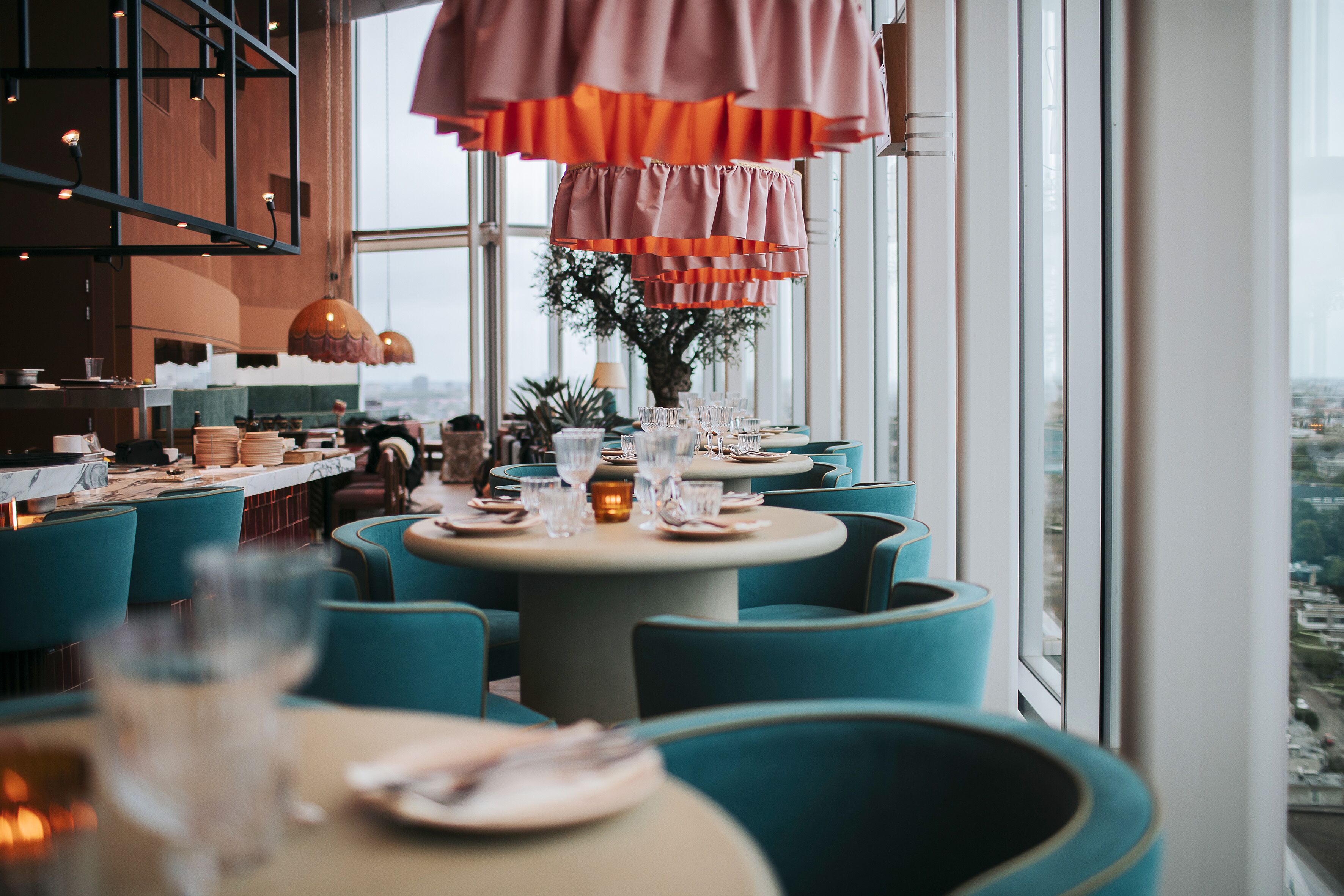 A restaurant with round tables, teal chairs, and large windows offering a city view.