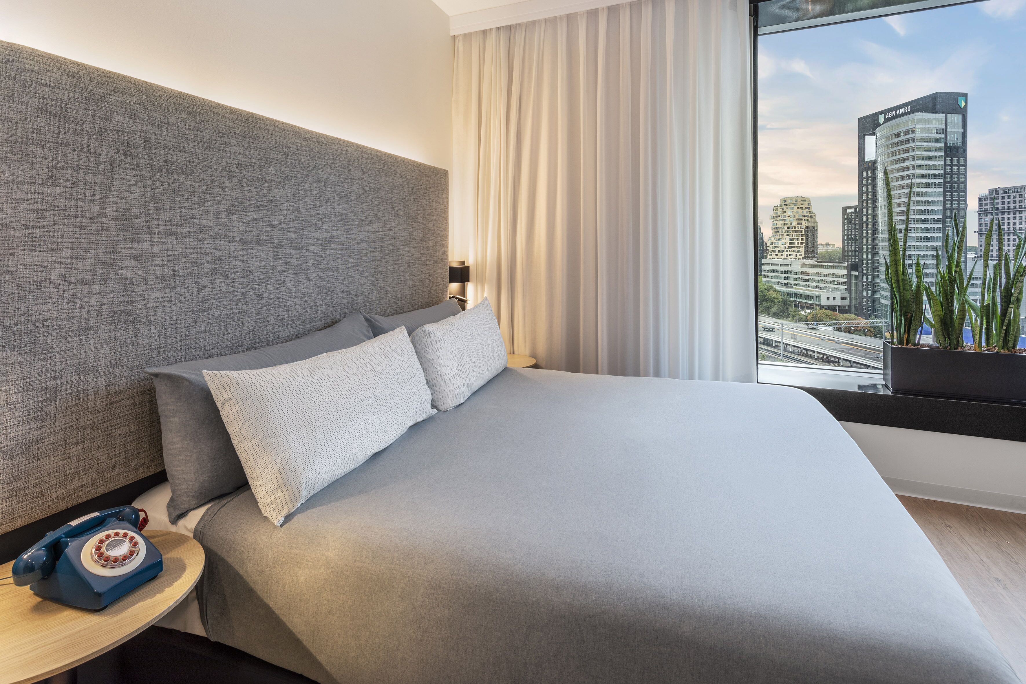 A modern hotel room with a large bed, a telephone on a side table, and a view of city buildings through the window.