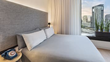 A modern hotel room with a large bed, a telephone on a side table, and a view of city buildings through the window.