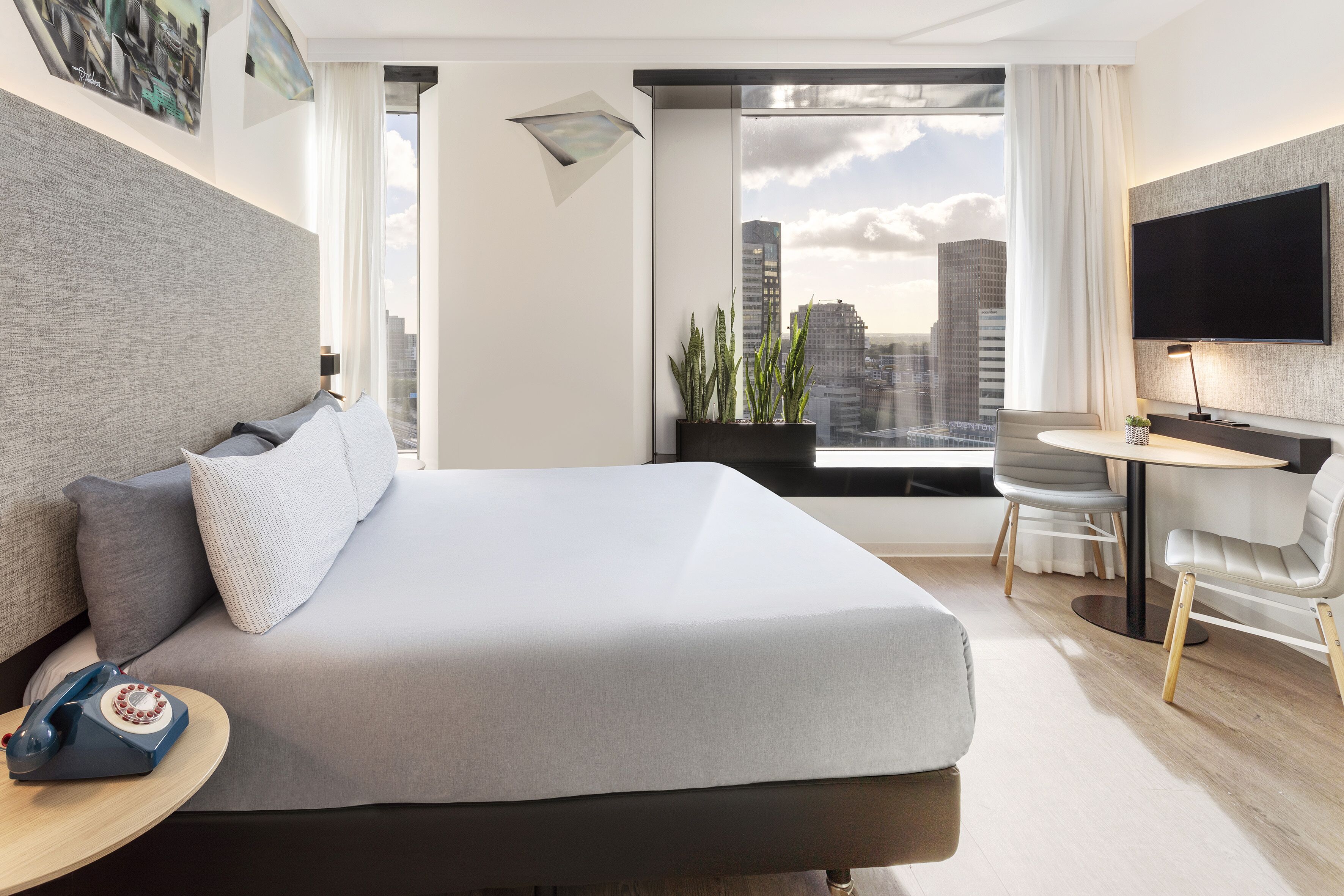 A modern hotel room with a large bed, a desk with a chair, a flat-screen TV, and a view of the cityscape.