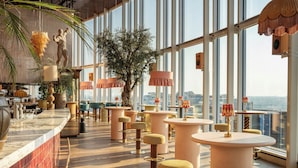 A modern bar area with round tables, bar stools, and large windows offering a city view.