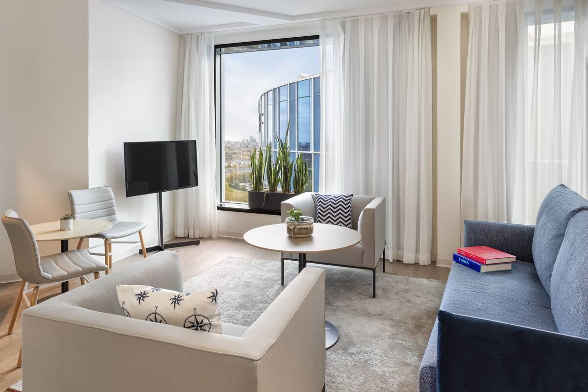 Uma sala de estar moderna com um sofá, uma mesa de centro, uma televisão e vista para o horizonte urbano através de grandes janelas.