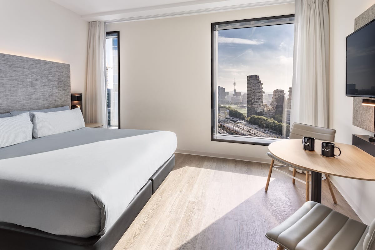 Quarto de hotel moderno com uma cama grande, uma mesa redonda com duas chávenas, uma televisão e vista para a cidade.