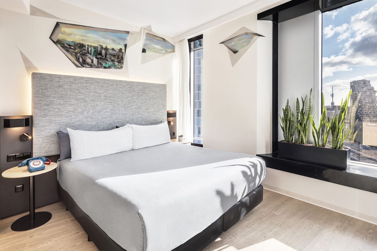 Quarto de hotel moderno com uma cama grande, uma pintura de paisagem urbana e uma janela com vista para prédios e uma torre.