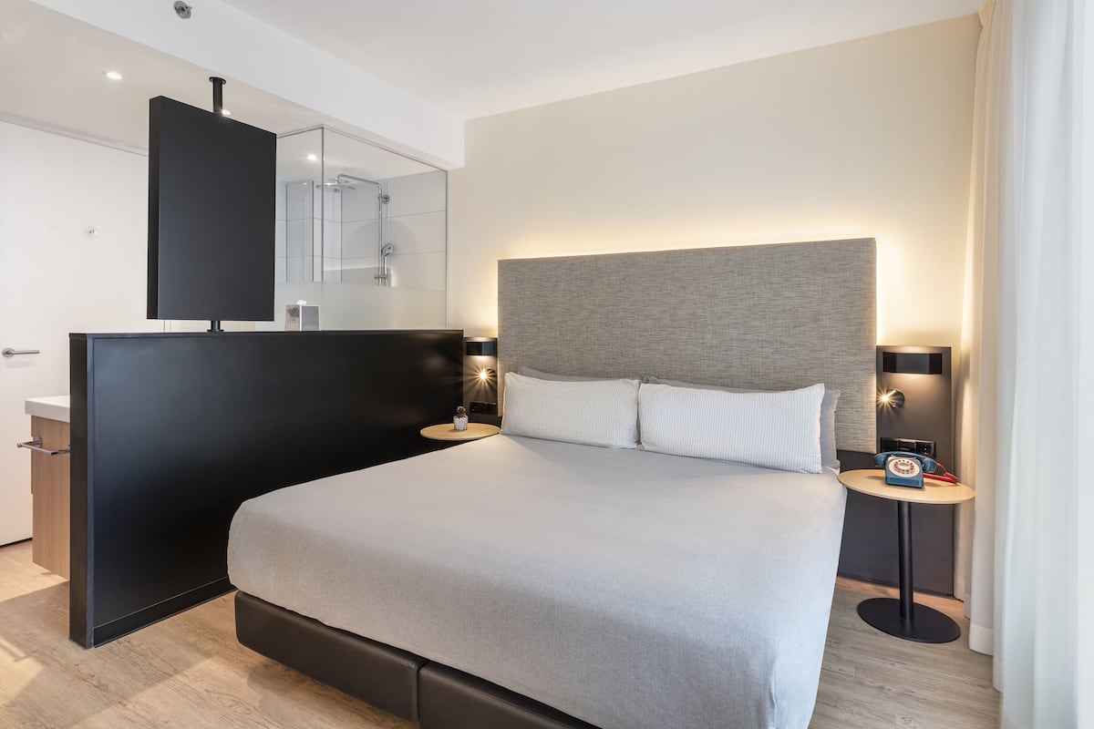 Quarto de hotel moderno com uma cama grande, uma mesa de cabeceira com abajur, um espelho e uma pequena mesa com telefone.