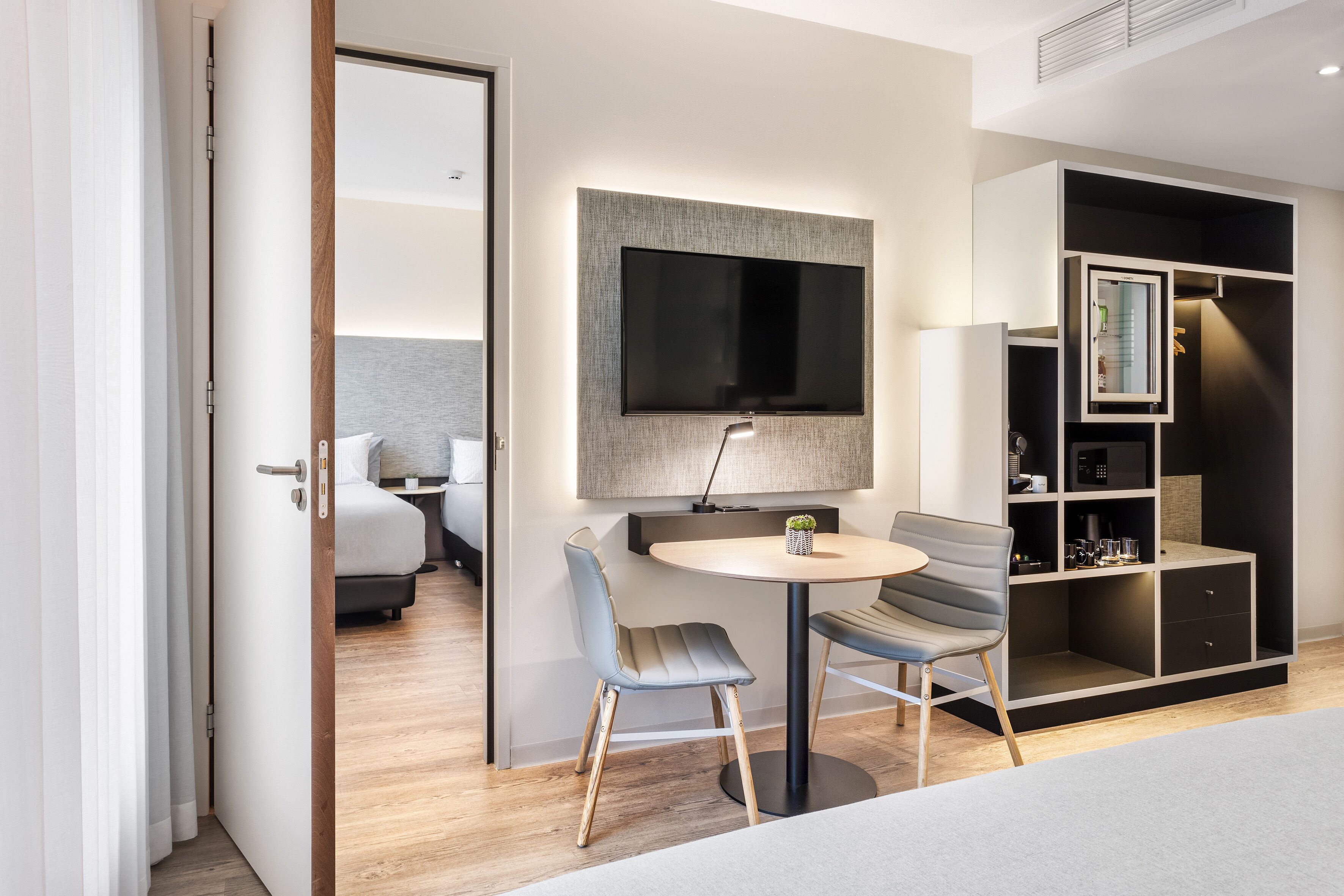 A modern hotel room with a bed, a TV mounted on the wall, a round table with two chairs, and a built-in wardrobe.