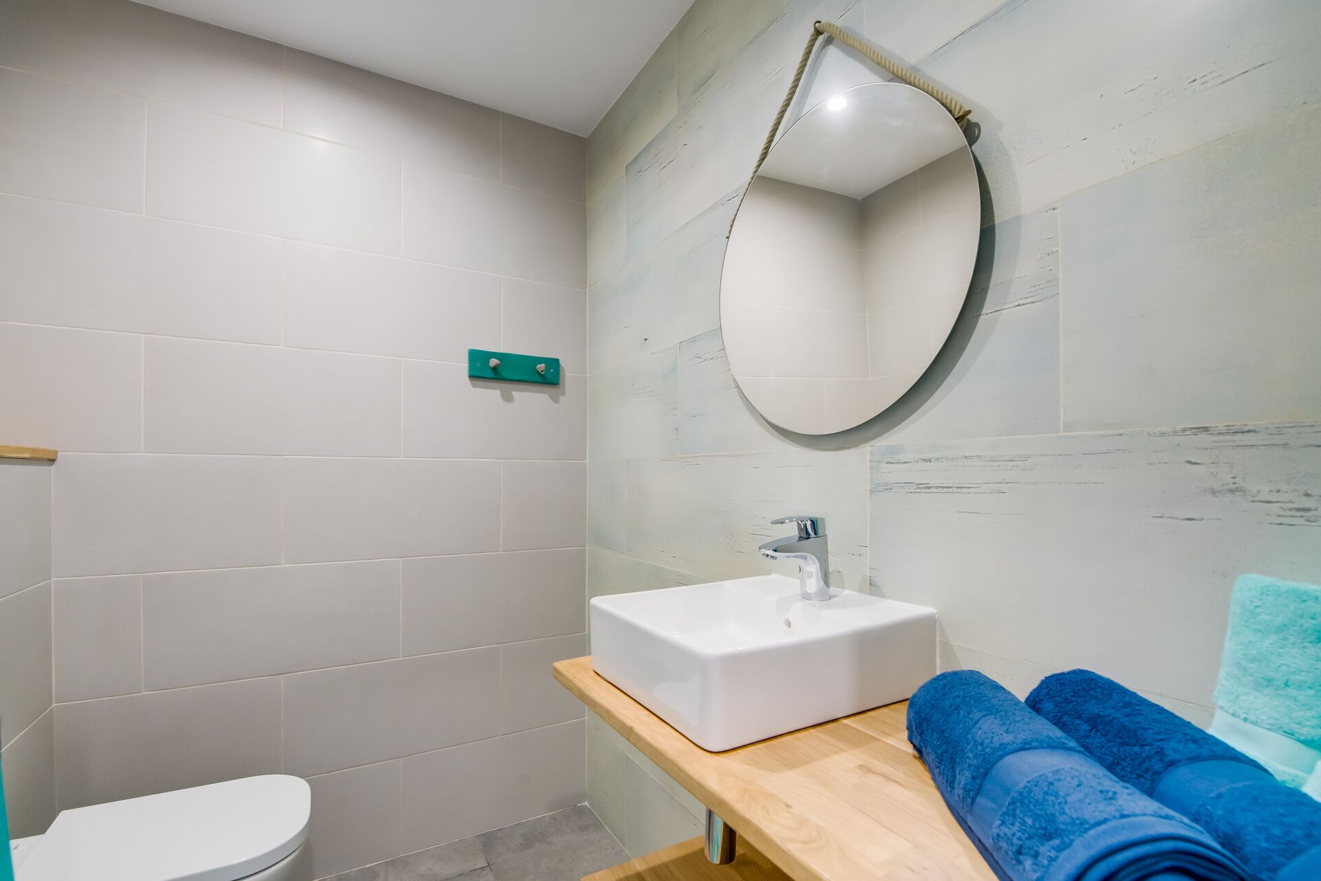 Un baño con inodoro blanco, un lavabo rectangular, un mueble de tocador de madera y un espejo redondo.