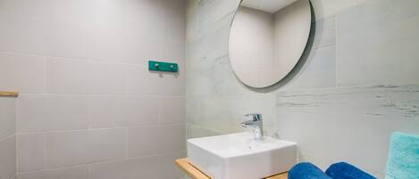 Un baño con inodoro blanco, un lavabo rectangular, un mueble de tocador de madera y un espejo redondo.