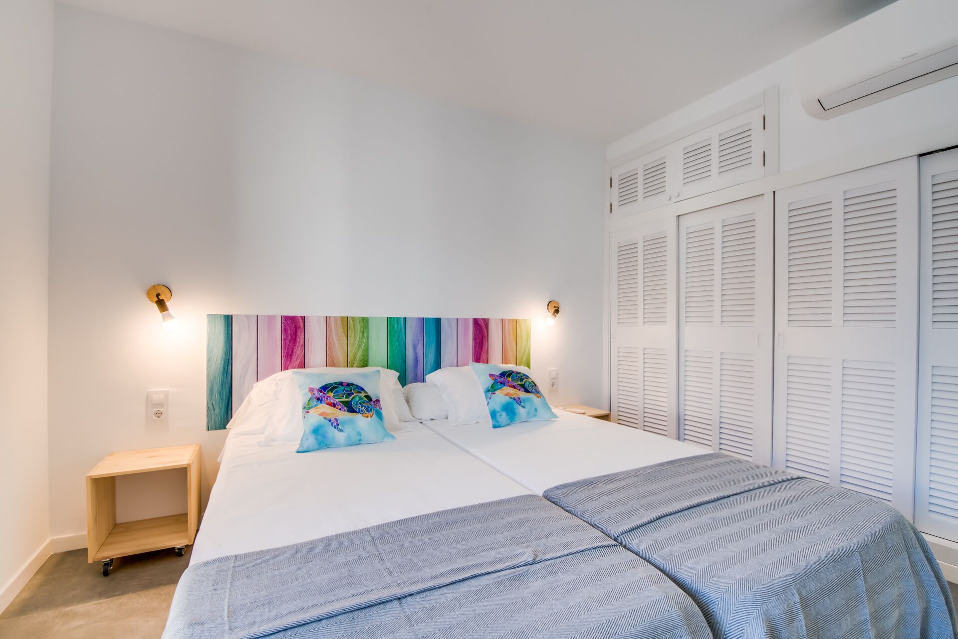 Une chambre à coucher comprenant un lit, deux tables de chevet, une lampe fixée au mur, une œuvre d’art colorée au-dessus du lit et une armoire à portes à persiennes.