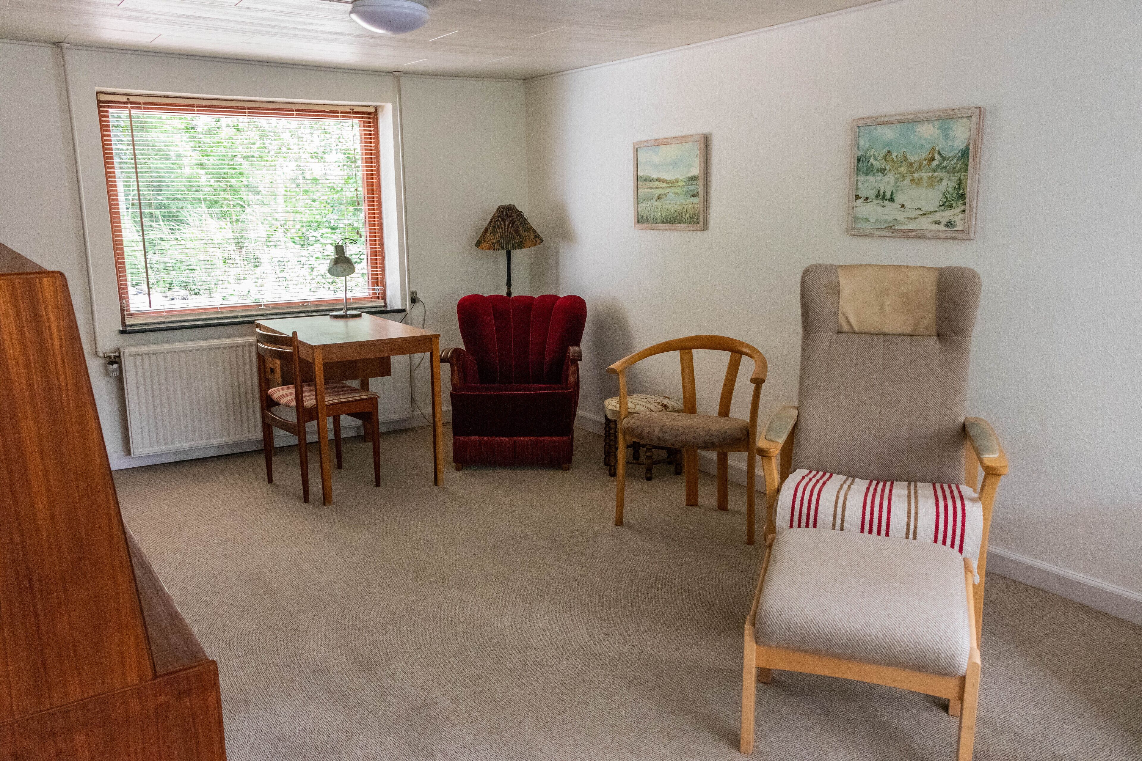 A room with a wooden table, two chairs, a red velvet chair, a lamp, and two paintings on the wall.