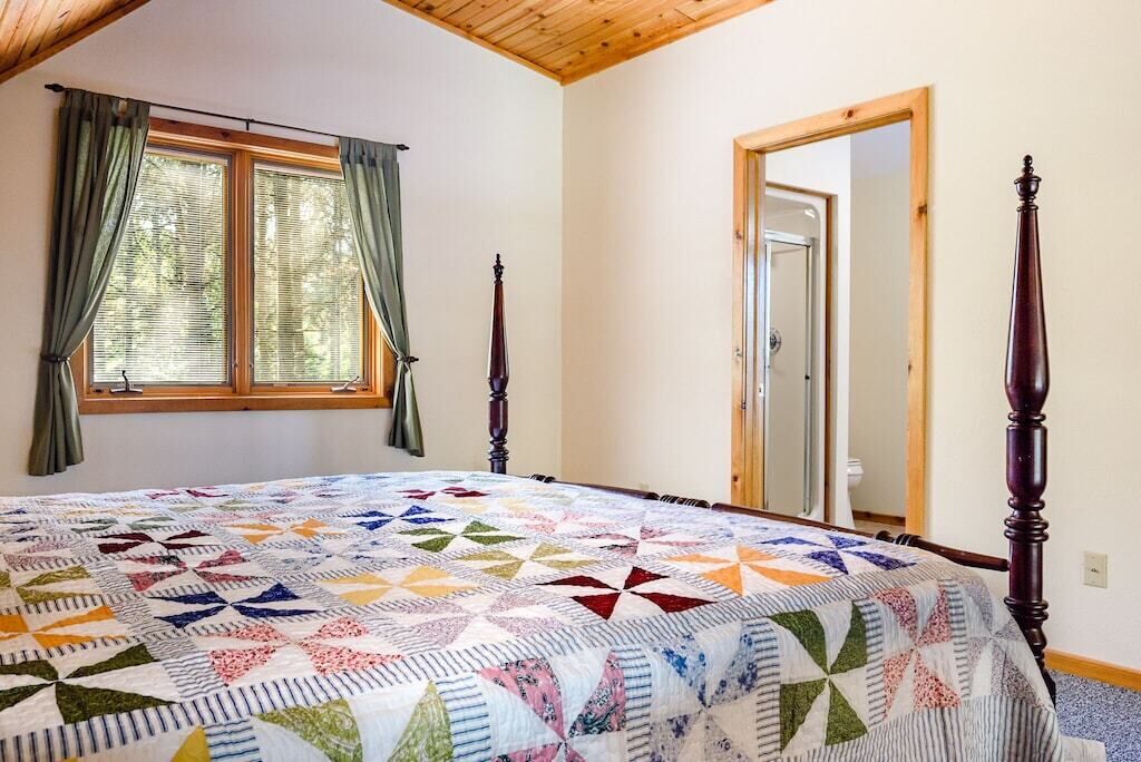 A bedroom with a large bed, a wooden ceiling, a window with curtains, and a mirror on the wall.