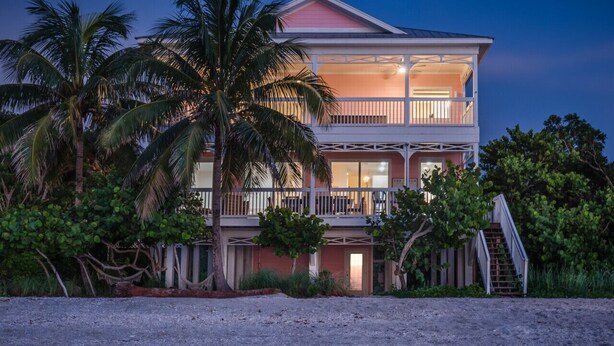 A two-story beachfront house with a balcony, palm trees, and a staircase leading to the entrance. - Pink Flamingo Beach House Private Beachfront Paradise with Pool Sleeps 12 (North Captiva)