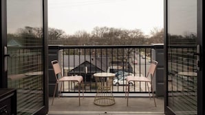 A balcony with two chairs and a table, a metal railing, and a view of a parking lot and trees. - Coffee Shops, Restaurants, Bars, Salons and Boutiques, just steps away. (Nashville)