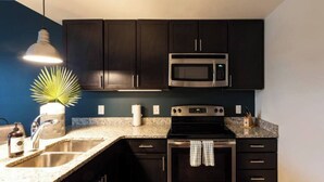 A modern kitchen with dark cabinets, a stainless steel sink, a microwave, an oven, and a granite countertop. - Coffee Shops, Restaurants, Bars, Salons and Boutiques, just steps away. (Nashville)