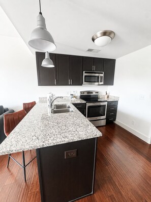 A modern kitchen with dark cabinets, stainless steel appliances, a granite countertop, and a pendant light.