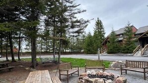Un espace extérieur aménagé avec des bancs en bois et des tables de pique-nique, entouré d’arbres et agrémenté d’un foyer en pierre.