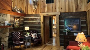 L’intérieur d’une cabane en rondins, avec une cheminée, des fauteuils en cuir, un plafond en bois et une tête d’animal empaillée accrochée au mur.