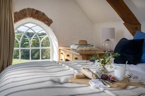 A cozy bedroom with a large window, a wooden dresser, a bed with a striped blanket, a vase with flowers, a mug, and a loaf of bread on a cutting board.
