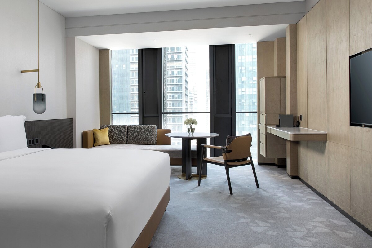 A hotel room with a large bed, a small table with a chair, a TV, and a view of tall buildings through the windows.