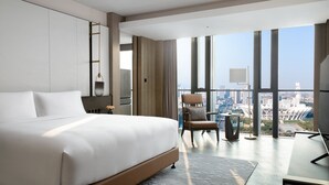 Une chambre d’hôtel moderne dotée d’un grand lit, d’un coin salon avec un fauteuil et une table, et offrant une vue sur le paysage urbain grâce à de grandes fenêtres. On peut également admirer la vue sur la ville depuis la fenêtre.