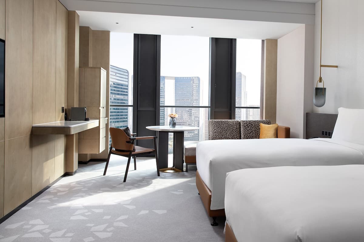 A hotel room with two beds, a desk, a chair, and a view of the cityscape.