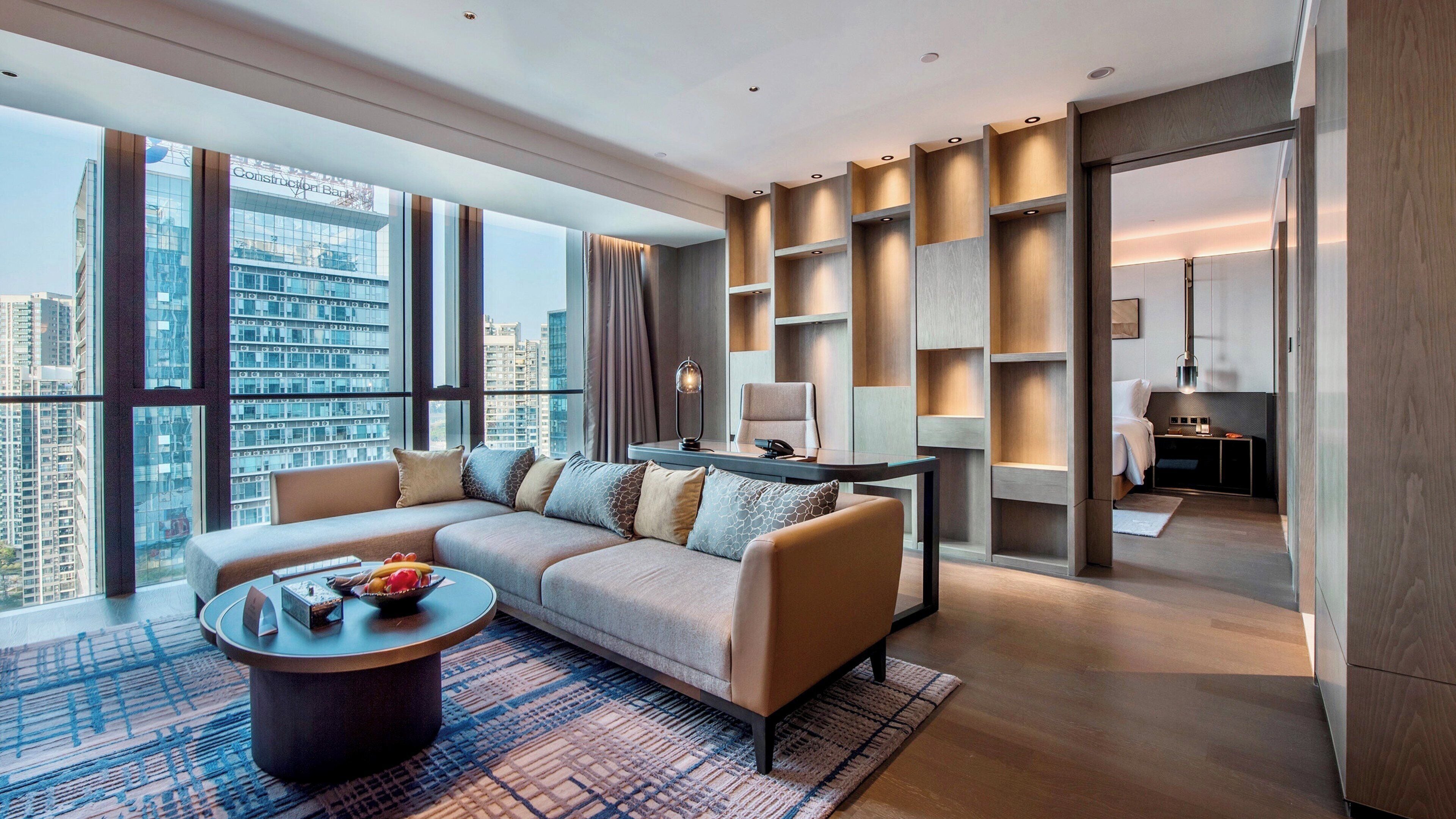 A modern hotel room with a large sofa, a coffee table, and a view of the cityscape.