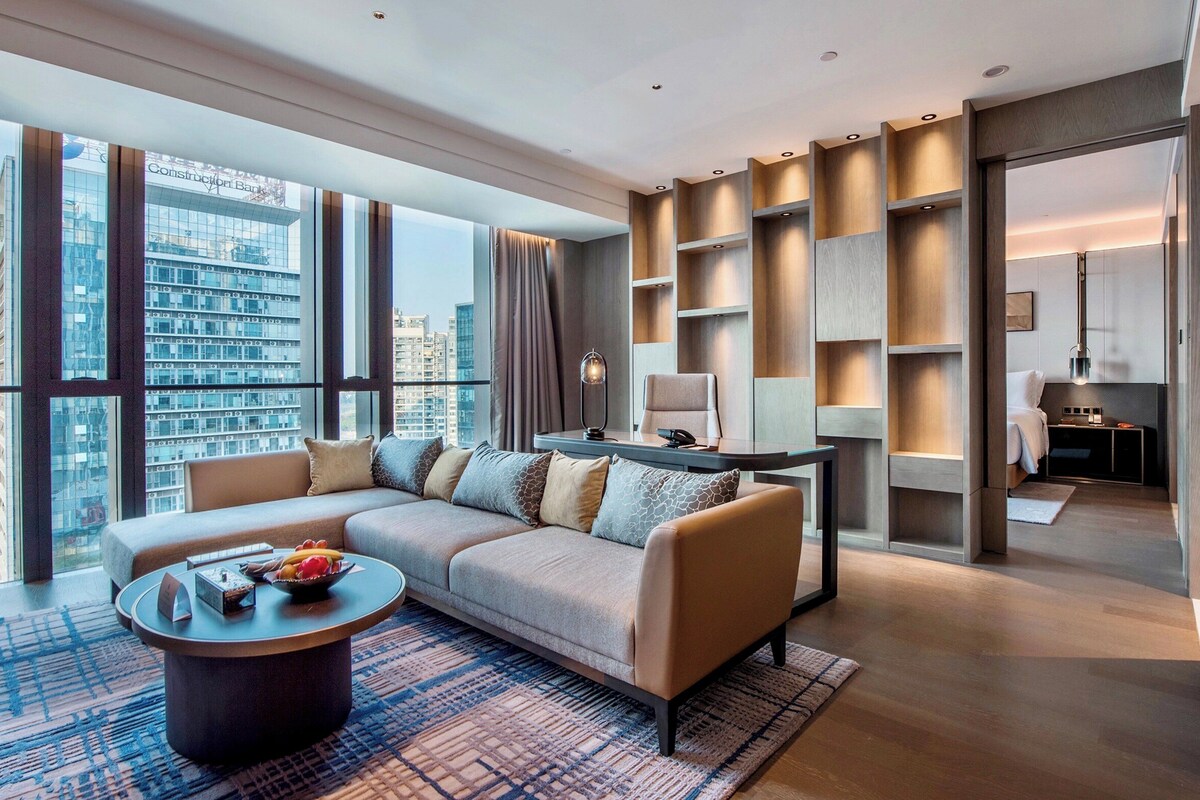 A modern hotel room with a large sofa, a coffee table, and a view of the cityscape.