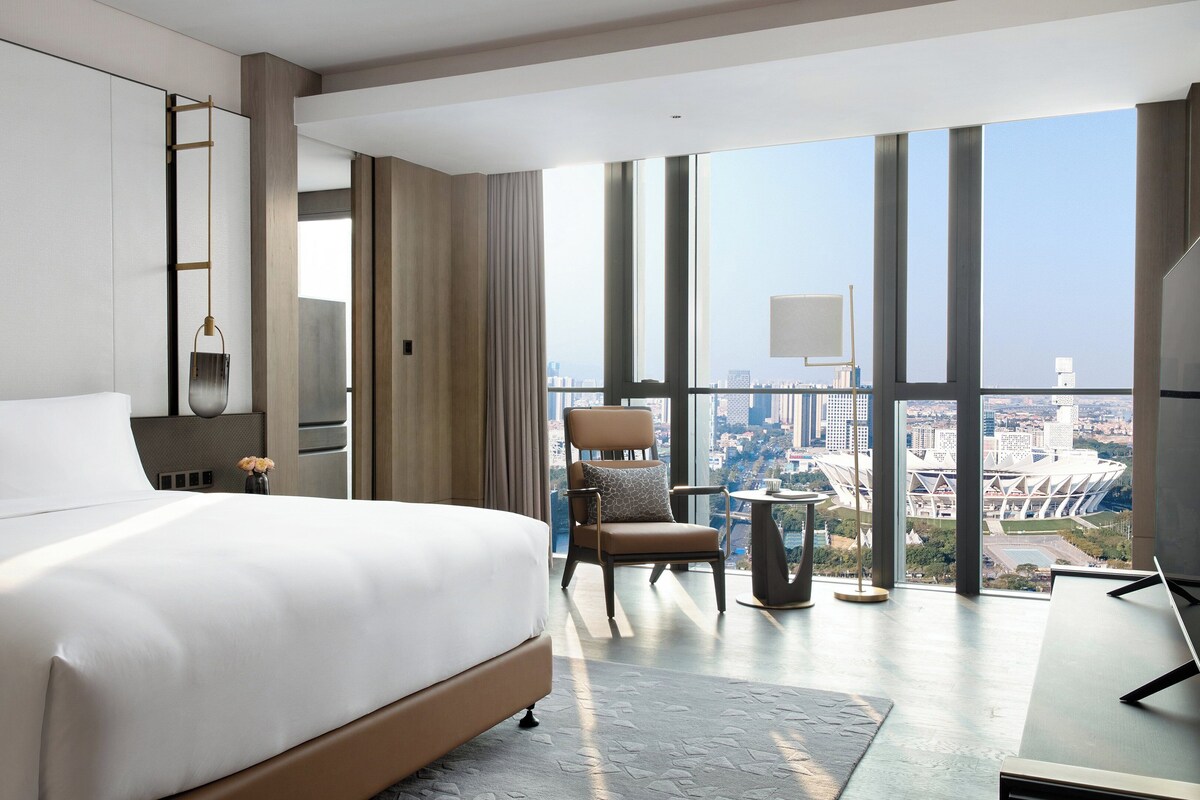 A modern hotel room with a large bed, a seating area, a view of the city, and a large window.