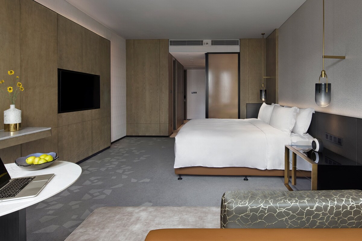A modern hotel room with a bed, a desk with a laptop, a bedside table, and a sofa.
