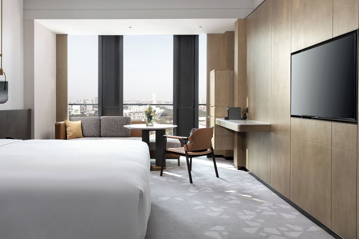 A hotel room with a large bed, a small table, a chair, a TV, and a view of the city.