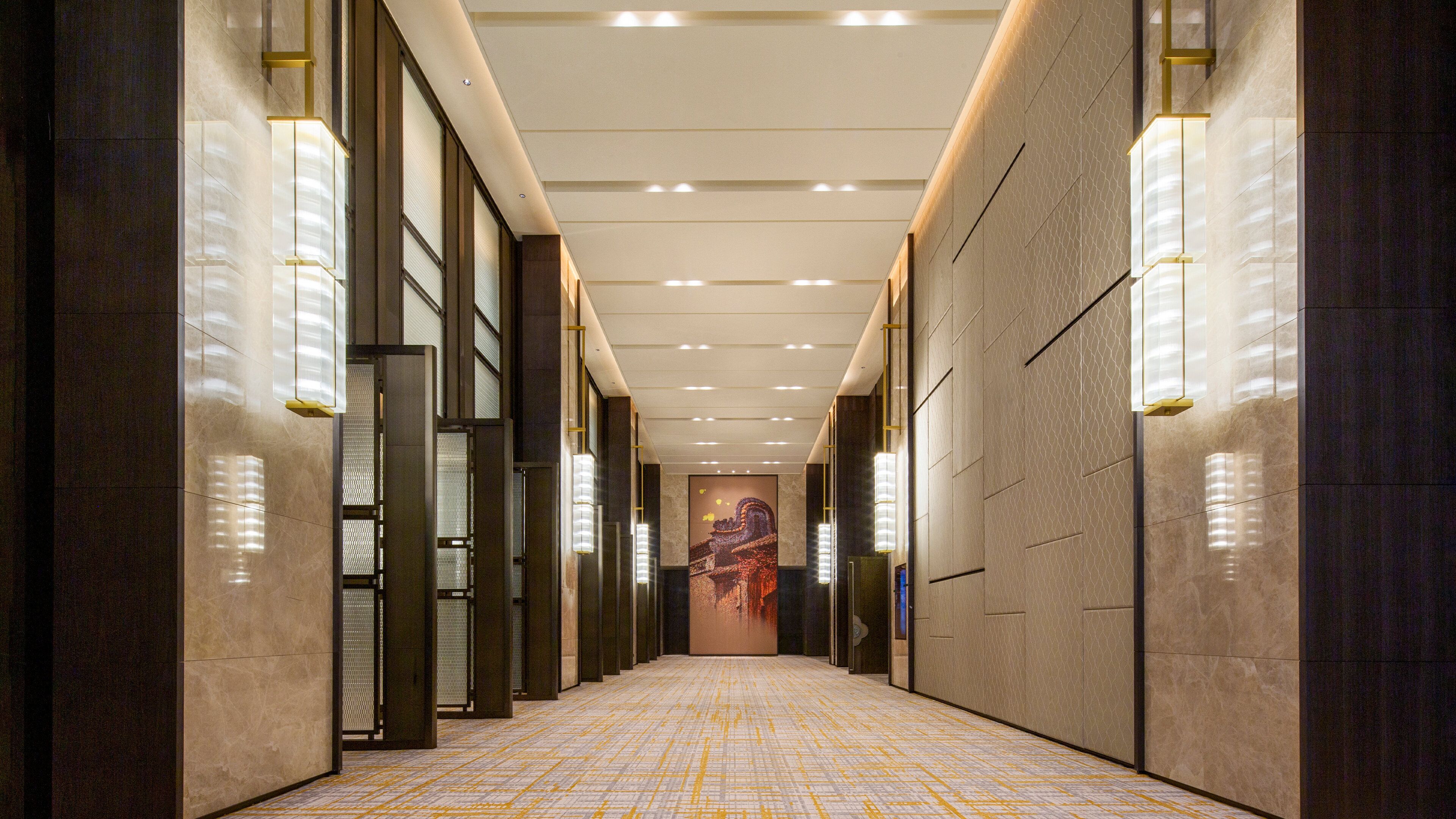 A long, narrow hallway with a patterned floor, recessed lighting, and a large wall-mounted artwork at the end.