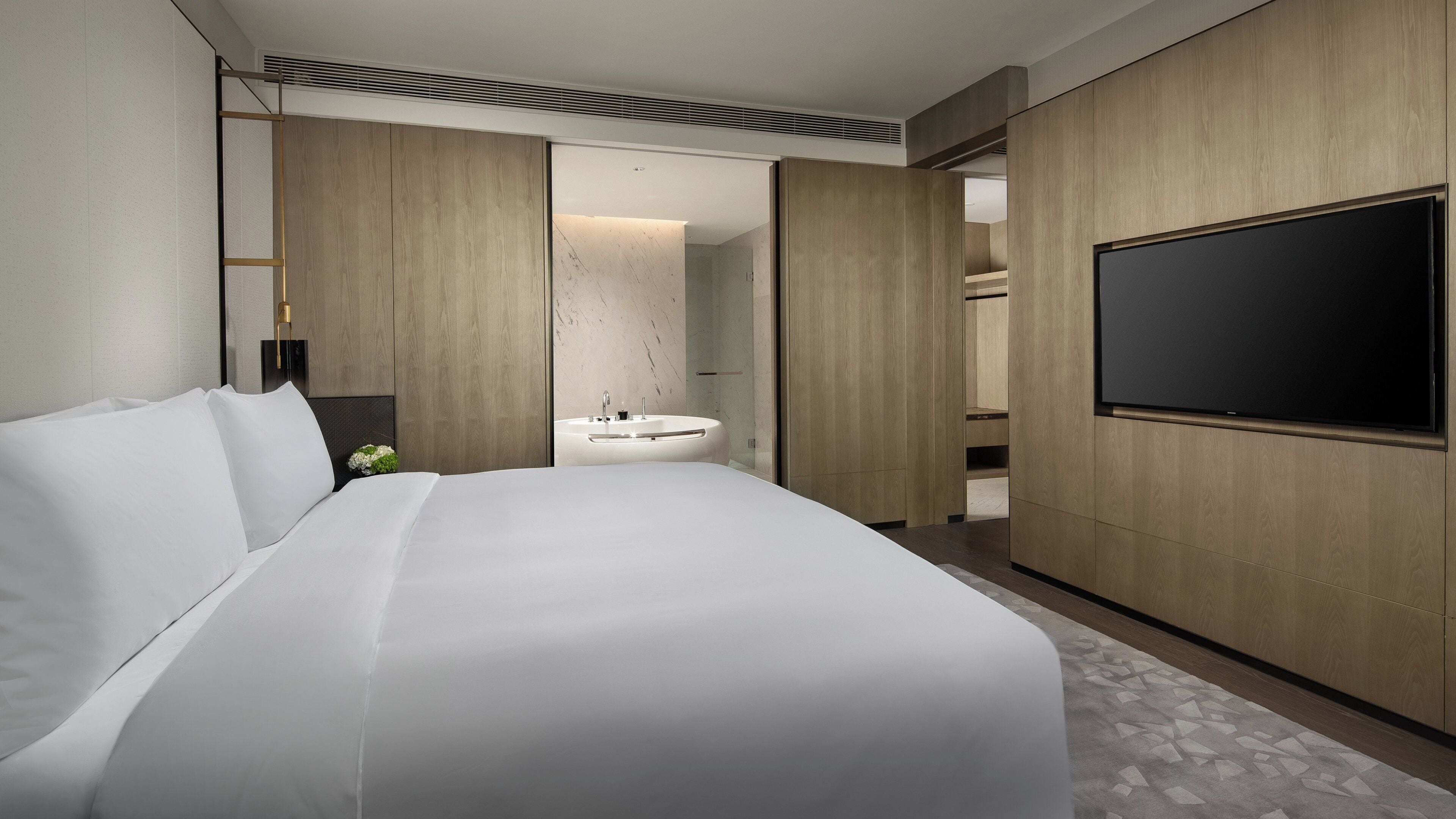 A modern hotel room with a large bed, a built-in TV, a bathroom with a sink, and a shower.