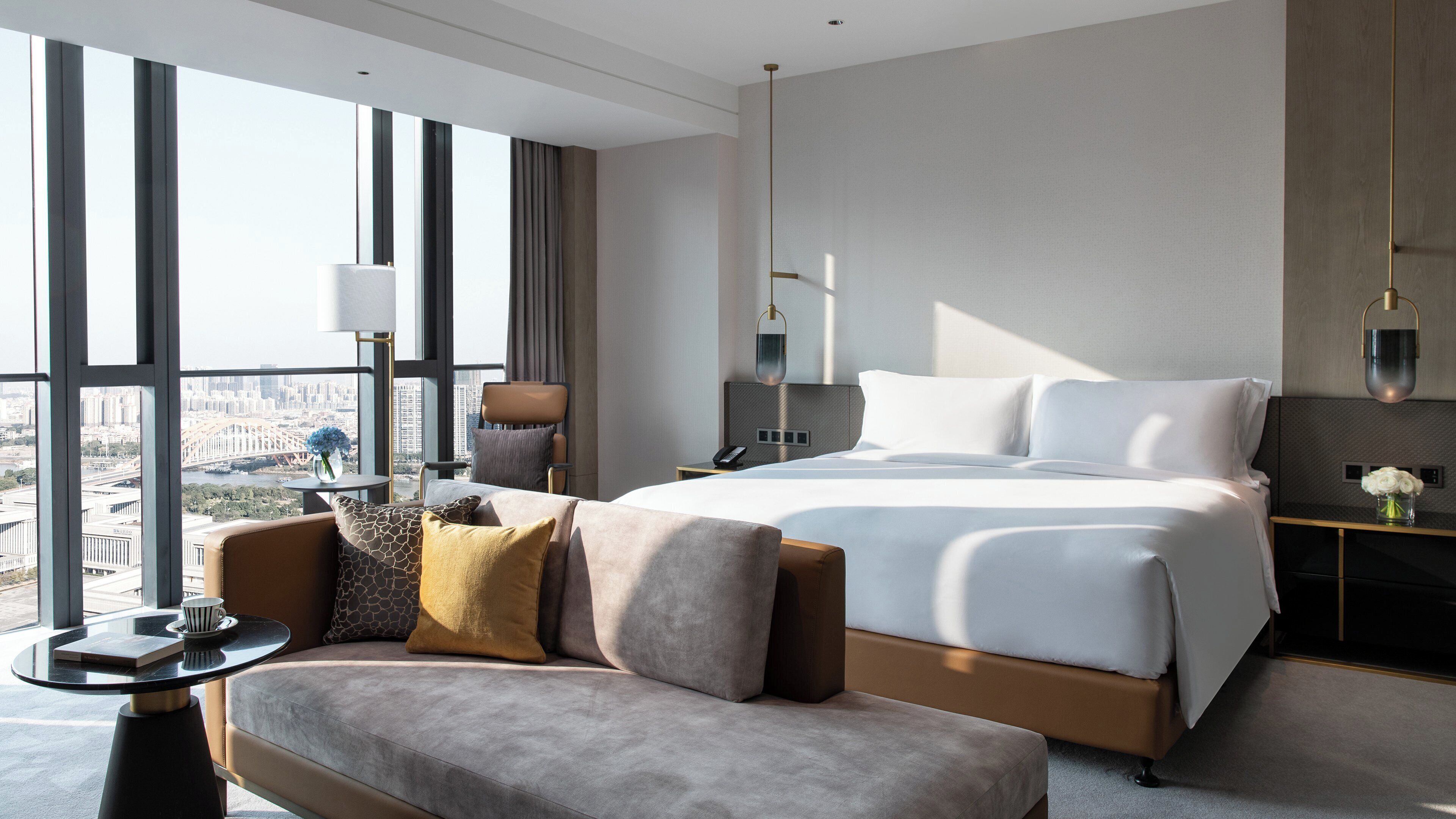 A modern hotel room with a large bed, a sofa, a small table, and a view of the city.