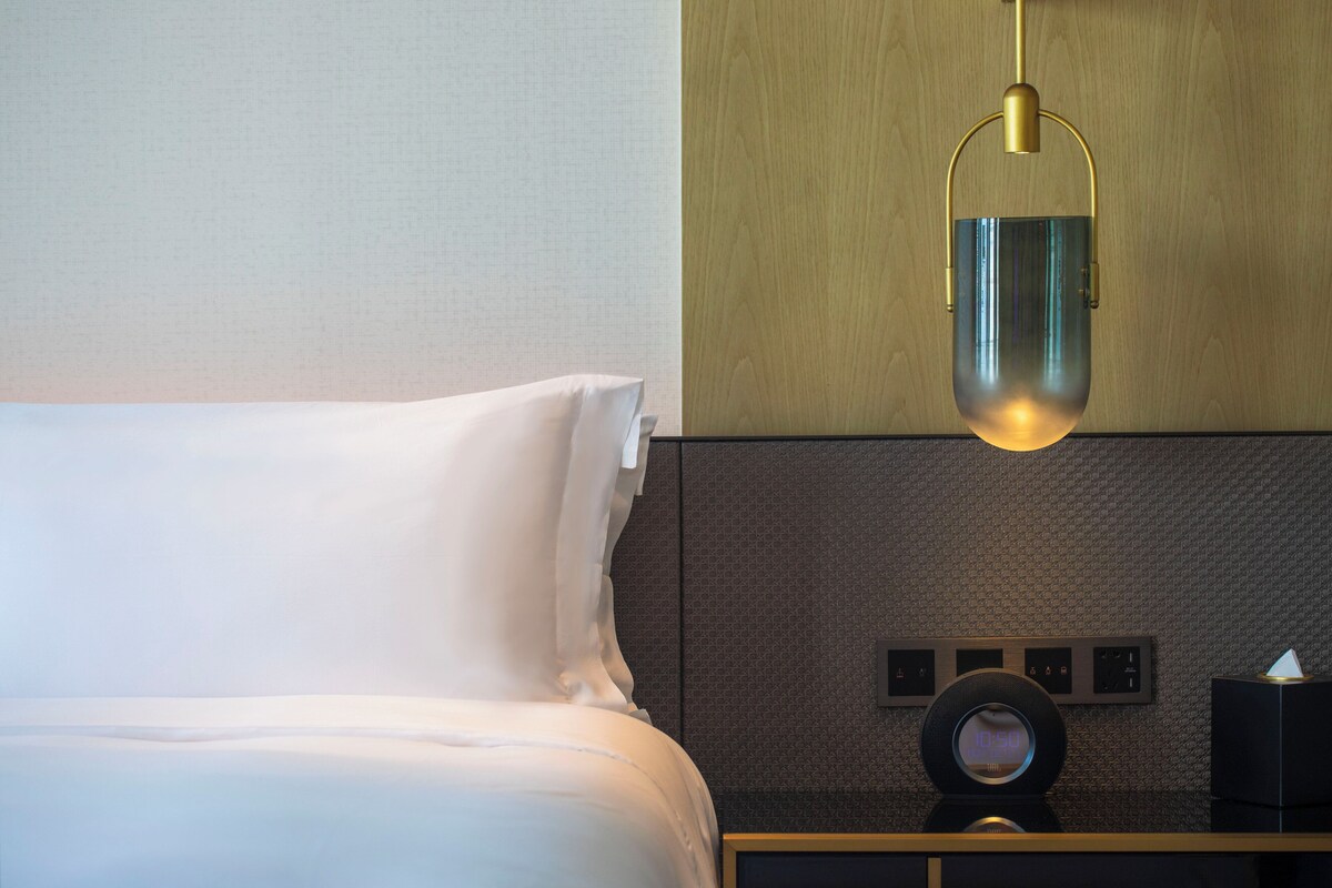 A hotel room with a bed, a bedside table with a clock, and a wall-mounted control panel.