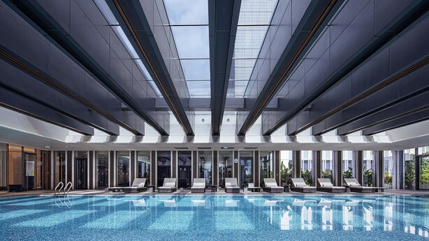 Une vaste piscine intérieure à l’eau d’un bleu cristallin, entourée de chaises longues et dotée d’une architecture moderne avec de grandes fenêtres et une verrière.