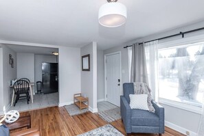 A modern living room with a grey sofa, a dining area with a table and chairs, a kitchen with a black refrigerator, and a large window with curtains. - Peaceful Views- 2BR Main Level Living w/Recliner and Adjustable Bed (Rochester)