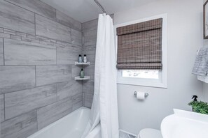 Une salle de bain moderne avec des murs recouverts de carreaux gris, une douche avec un rideau blanc, une fenêtre avec un store marron et un lavabo blanc agrémenté d'une plante.